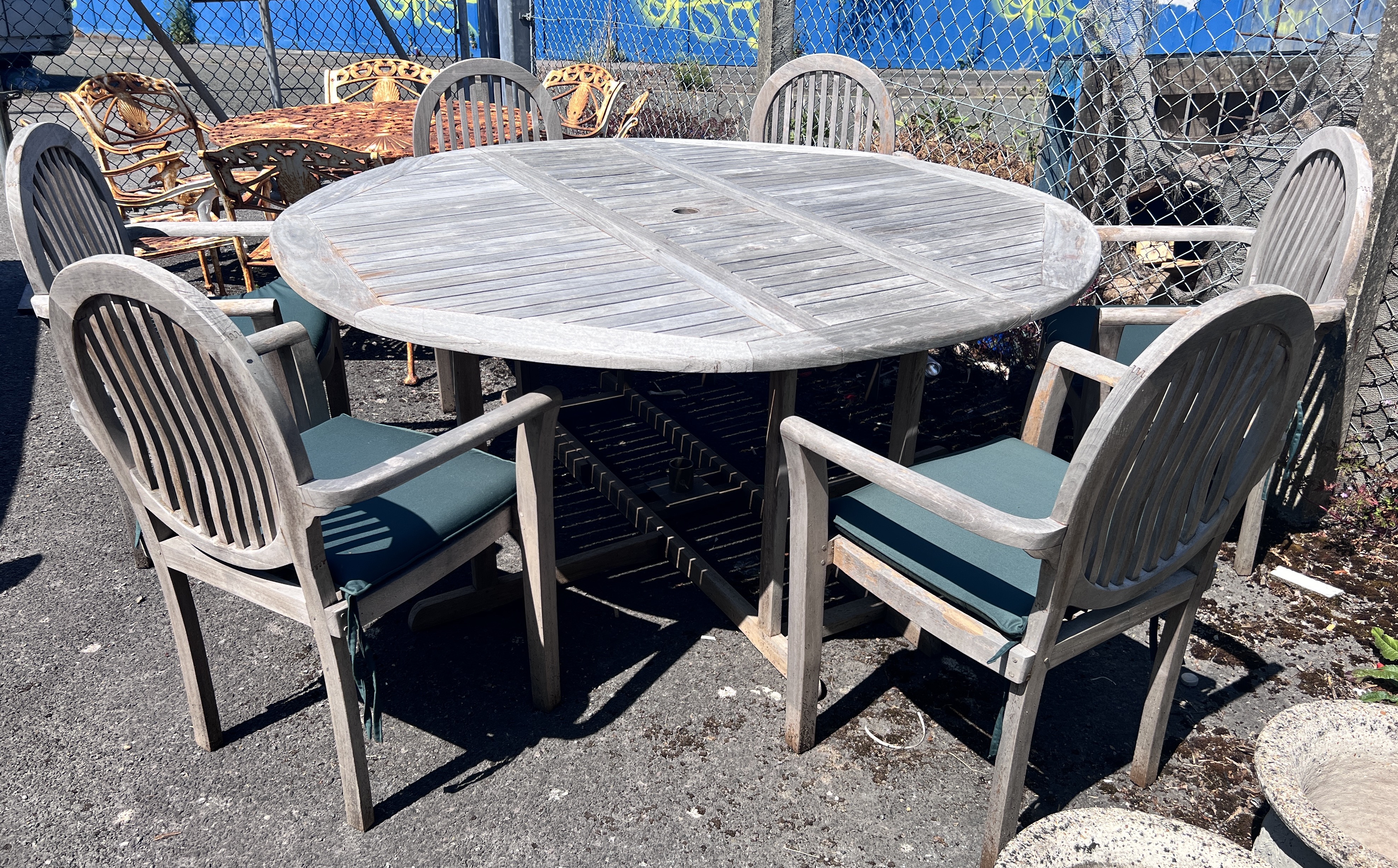 A circular weathered teak drop leaf garden table, 180cm extended, height 78cm, and six elbow chairs with seat cushions